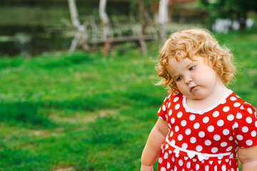 A young child wearing a bright red dress with white polka dots stands with a thoughtful expression in a grassy area The sun casts a warm light as she gazes down
