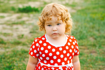 A young child with blonde curly hair gazes thoughtfully while wearing a bright red dress adorned with white polka dots The grassy background enhances the playful atmosphere of the moment