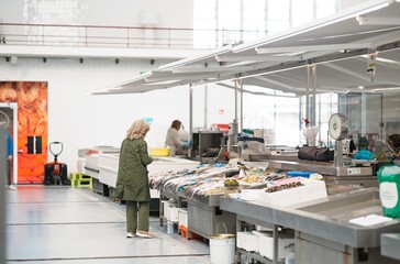 An older woman in a green coat carefully examines the selection of fresh fish at a stall inside the...