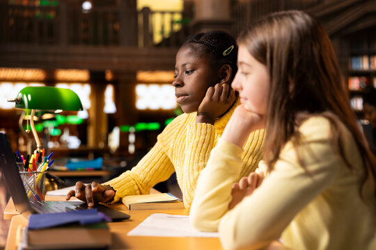 Two schoolgirls in the library participate at an online class webinar, using technology to connect with their teacher for an interactive virtual lesson. Remote courses via video call.