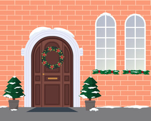 The front door to the house on the facade and two windows. Winter holidays - Christmas and New Year. Snow-covered trees and a door decorated with a Christmas wreath of fir branches. Festive atmosphere