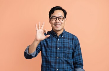 Smiling Asian man in blue plaid shirt gives OK gesture, confirming agreement or satisfaction. He wears glasses and stands against a plain peach background, suggesting confidence and positivity.