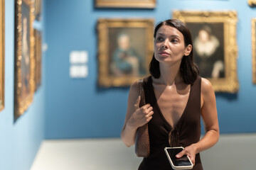 Female visitor with phone walks along painting exhibition pavilion. She inspects paintings,...