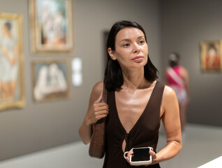 Museum girl visitor with phone in hands walk in exhibition halls of museum. Enjoy contemplation, be inspired by painting, get acquainted with art of other cultures, cultural heritage
