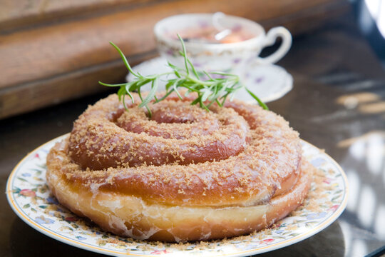 A close-up of a beautifully glazed spiral pastry topped with brown sugar and garnished with fresh rosemary. Served on a floral vintage plate with a teacup in the background.