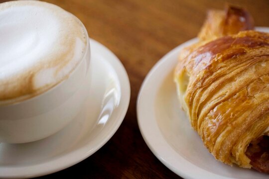 A warm and inviting image of a frothy cappuccino paired with a golden croissant on a wooden table, captured in natural light. A classic breakfast or caf&eacute; moment evoking comfort and indulgence.

