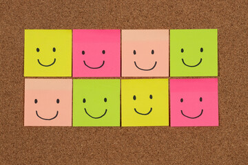 a group of multicolored sticky notes representing hapiness with a lot of happy faces over a cork...