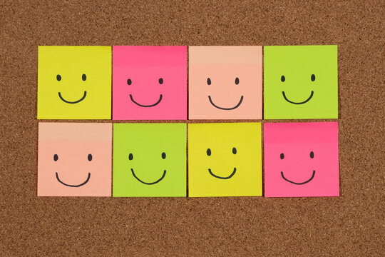 a group of multicolored sticky notes representing hapiness with a lot of happy faces over a cork background