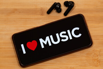a smartphone with i love music phrase at the screen and a wireless earphones over a wooden...