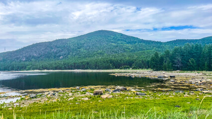  Mountain view over a calm coastal bay with rocky shore and pine forest under a partly cloudy sky. Peaceful wilderness scene in northern Russia.
