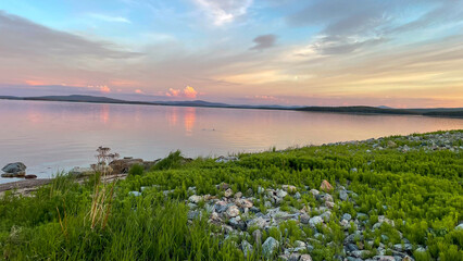  Coastal landscape at sunset with green grass, rocky shore, and calm waters reflecting the warm hues of the sky over the White Sea.