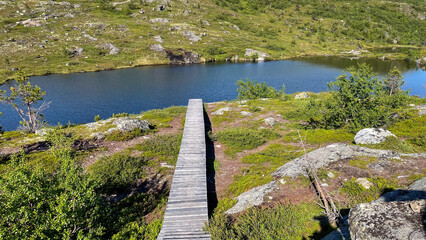 A narrow wooden footbridge crosses a calm lake on the Kola Peninsula in Murmansk Oblast, Russia, surrounded by rocky tundra and serene northern landscapes.