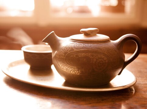 A beautifully crafted ceramic teapot with intricate carvings sits on a matching tray alongside a teacup. The warm, glowing sunlight creates a serene and cozy atmosphere.