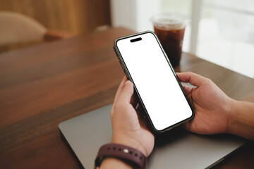 A person's hands holding a smartphone with a blank white screen over a laptop, with an iced coffee on the table. An ideal mobile app mockup.