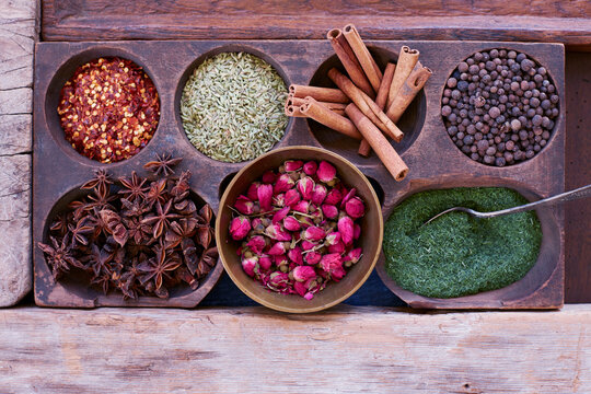 A vibrant overhead shot of assorted dried spices including cinnamon sticks, rosebuds, star anise, fennel seeds, black peppercorns, dried dill, and chili flakes displayed in a rustic wooden box.