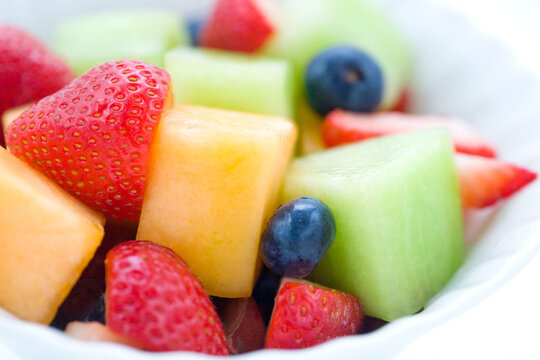 Macro shot of a vibrant and juicy fruit salad featuring strawberries, blueberries, cantaloupe, and honeydew melon in a white bowl.