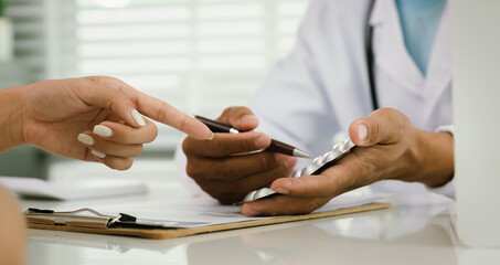 A doctor consults with a patient, explaining medical documents at a desk with a clipboard, laptop,...