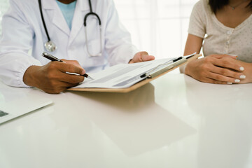 A doctor consults with a patient, explaining medical documents at a desk with a clipboard, laptop,...