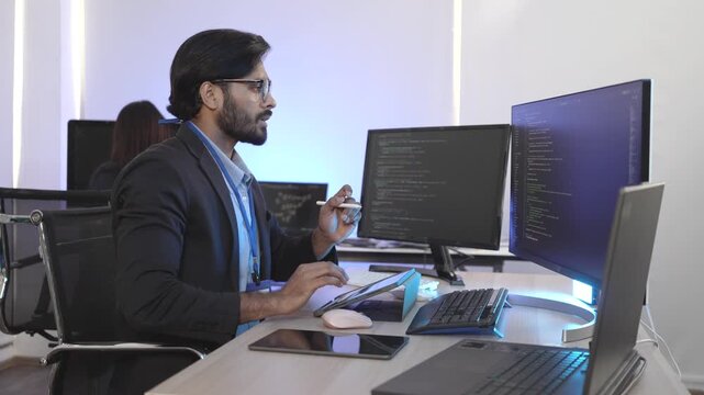 A focused Indian software engineer working on a laptop at his multi-monitor workstation in a modern tech office. Concept for deep work, programming, and data analysis.