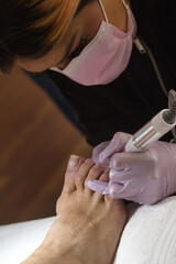 Close-up of a pedicurist wearing pink gloves and face mask removing gel polish from toenails using...