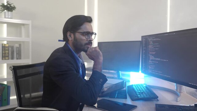 A focused Indian software engineer working on a laptop at his multi-monitor workstation in a modern tech office. Concept for deep work, programming, and data analysis.