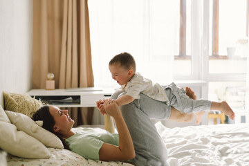 Home portrait of a baby boy with mother on the bed relaxing and playing in the bed at the weekend together, lazy morning, warm and cozy scene. Mom holding and kissing her child. Mother day concept