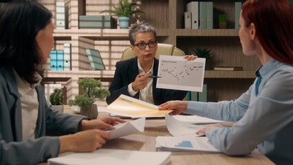 Caucasian team leader showing charts pointing graphs coworkers work business analyzing corporate discuss statistics diverse women businesswomen paperwork office boardroom planning data group documents