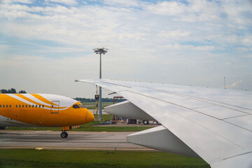 Aviation image capturing the wing of a commercial airplane under a cloudy sky, alongside a yellow airplane prepared on the runway. The detailed texture suggests modern aviation technology and travel.