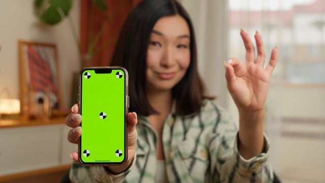 Asian Korean woman showing mobile phone green screen at camera smartphone app gesture ok okay good female at home mock up chroma key indoor Chinese Japanese girl recommend online social media display
