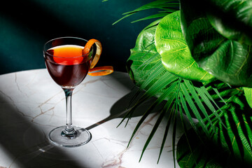 Elegant cocktail served in a glass with garnished orange peel on a marble table surrounded by lush...