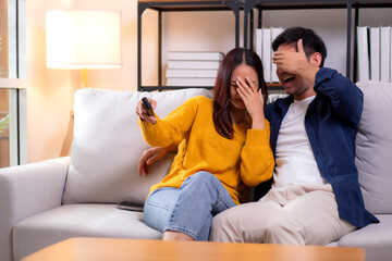 Young asian couple watching a scary or thrilling TV show in living room at home with emotion shock...