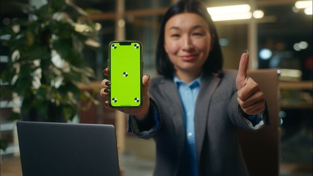 Asian Korean business woman businesswoman holding mobile phone showing smartphone green screen chroma key display Chinese Japanese female girl smiling thumb up advertise app recommend office workplace