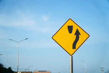 A vibrant yellow road sign indicating lane merging ahead, set against a bright sky backdrop. This image highlights roadway safety and infrastructural elements on a sunny day.A vibrant yellow road sign