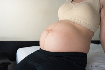 Pregnant asian woman sitting on bed in a softly lit room with natural light streaming through the...