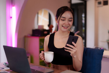 Asian woman freelancer digital nomad working online using smartphone drinking coffee at modern...