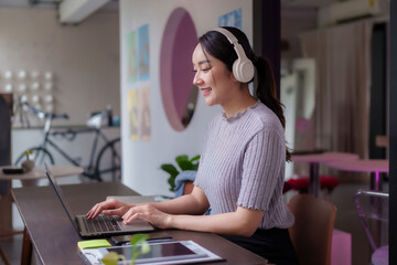 Freelance Asian adult woman working laptop headphones smiling coworking space digital nomad remote...
