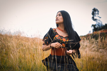 Indigenous woman wearing traditional, embroidered clothing and accessories poses in a field of tall