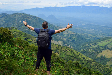 Male hiker with a backpack seen from behind, standing on a mountain summit and looking at the spectacular view of the green valley and landscape during an adventure travel trip in the Andes, Colombia.