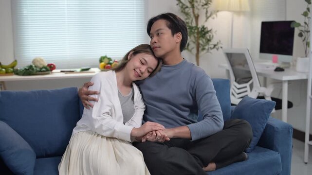man and a woman are sitting on a couch, holding hands and looking at each other. The woman is wearing a white shirt and the man is wearing a blue shirt. Scene is warm and affectionate
