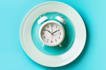 Empty plate with alarm clock on blue background, intermittent fasting concept,