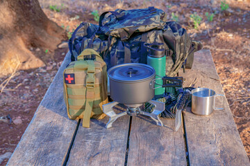 Essential camping gear including backpacks, cooking utensils, first aid kit, and water bottle arranged on a wooden table in nature