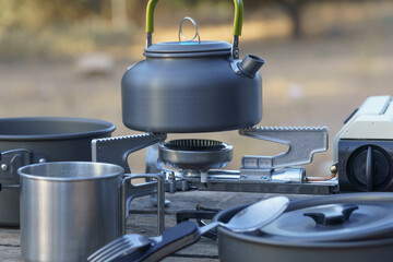 Camping kettle boiling on portable gas stove, making a hot drink outdoors, surrounded by camping cookware