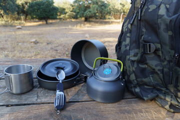 Camping cookware set and backpack on wooden table in nature, ready for outdoor cooking