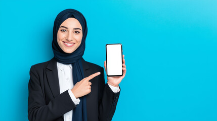 Smiling Muslim businesswoman wearing hijab pointing at a smartphone with blank screen. Ideal for app mockups, tech promotion, and diverse professional business concepts.