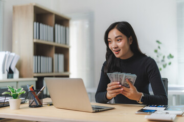 Excited businesswoman holding cash while looking at laptop in office