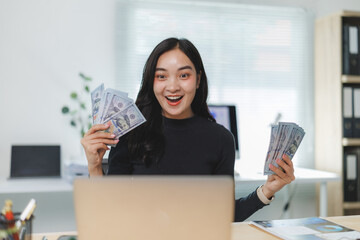 Excited businesswoman holding us dollar banknotes in office, making money online