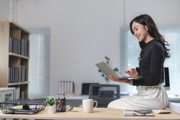 Young asian businesswoman using laptop and sitting on desk in modern office