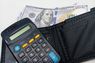 A black foid wallet contains several US $100 bills, with a dark-colored calculator partially...