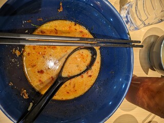 Top-down view of an empty blue ramen bowl with leftover spicy broth, black chopsticks, and a soup spoon. Surrounding items include a water bottle, a cup, and a glass of i