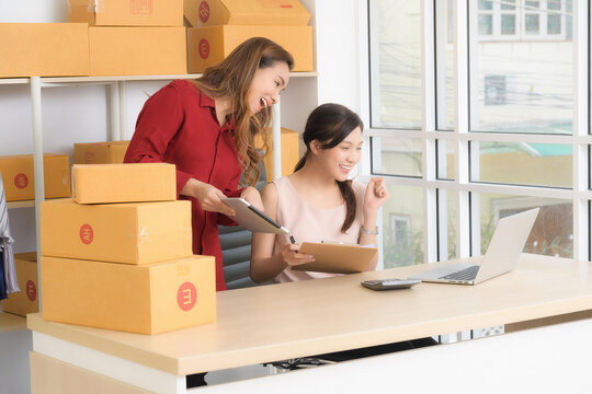 Two smiling Asian women, business partners, happily discuss operations and review documents for their online store in a bright home office setting.